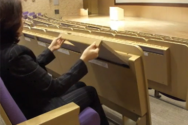 A lady in a conference room. She is folding a writing tablet into a housing in the chair in front.